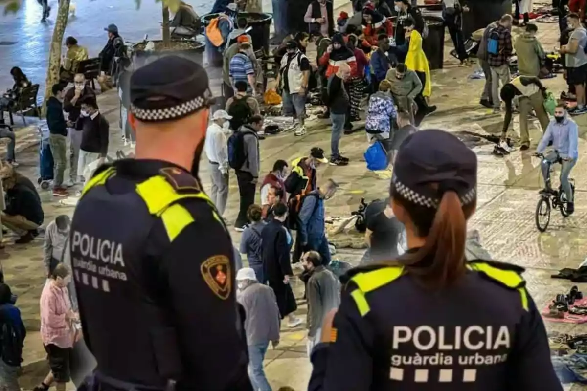 Dos agentes de la policía observan una concurrida plaza donde hay muchas personas reunidas, algunas caminando y otras en bicicleta.