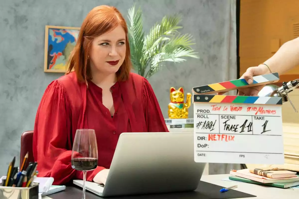 Elísabet Benavent pelirroja con blusa roja sentada frente a una laptop en un escritorio con una copa de vino, lápices y una claqueta de grabación, al fondo una planta, un cuadro colorido y una figura de gato dorado en Toda la verdad de mis mentiras.
