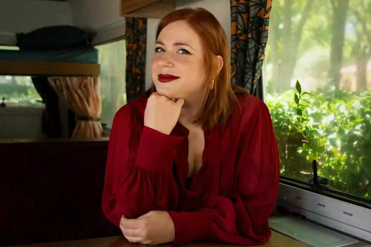 Elísabet Benavent con blusa roja sentada junto a una ventana en el interior de una caravana, apoyando el mentón en su mano y sonriendo suavemente en Toda la verdad de mis mentiras. Elísabet Benavent con blusa roja sentada junto a una ventana en el interior de una caravana, apoyando el mentón en su mano y sonriendo suavemente en Toda la verdad de mis mentiras.
