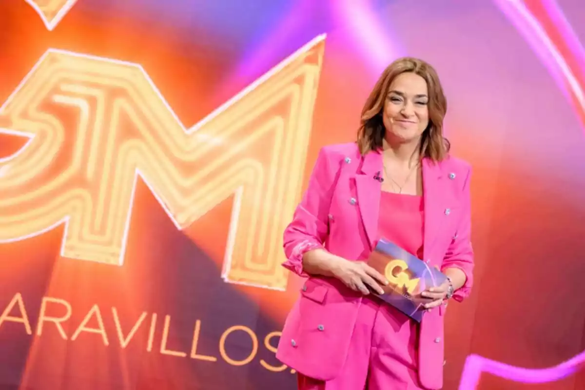 Toñi Moreno con traje rosa sosteniendo una tarjeta en un escenario con fondo colorido en Gente Maravillosa.