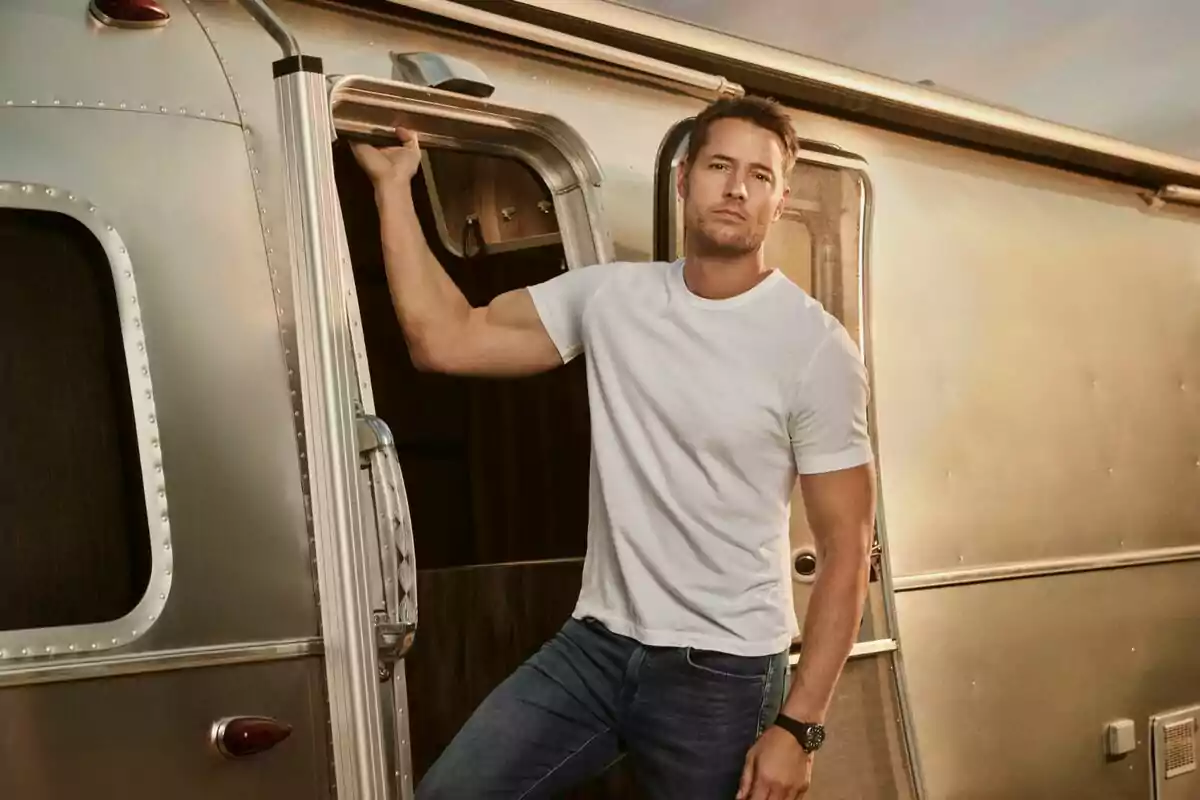 Justin Hartley de pie junto a la puerta de una casa rodante plateada vestido con camiseta blanca y jeans azules en la serie Tracker Justin Hartley de pie junto a la puerta de una casa rodante plateada vestido con camiseta blanca y jeans azules en la serie Tracker