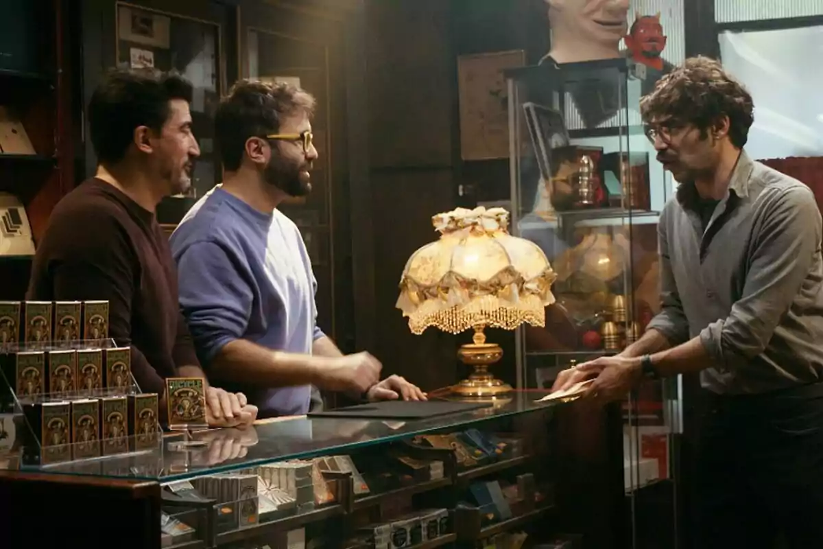 Pere Rafart, David Verdaguer y una tercera persona conversan en una tienda de magia, con una lámpara decorativa en el mostrador y estantes llenos de objetos curiosos en Tria una carta. Pere Rafart, David Verdaguer y una tercera persona conversan en una tienda de magia, con una lámpara decorativa en el mostrador y estantes llenos de objetos curiosos en Tria una carta.