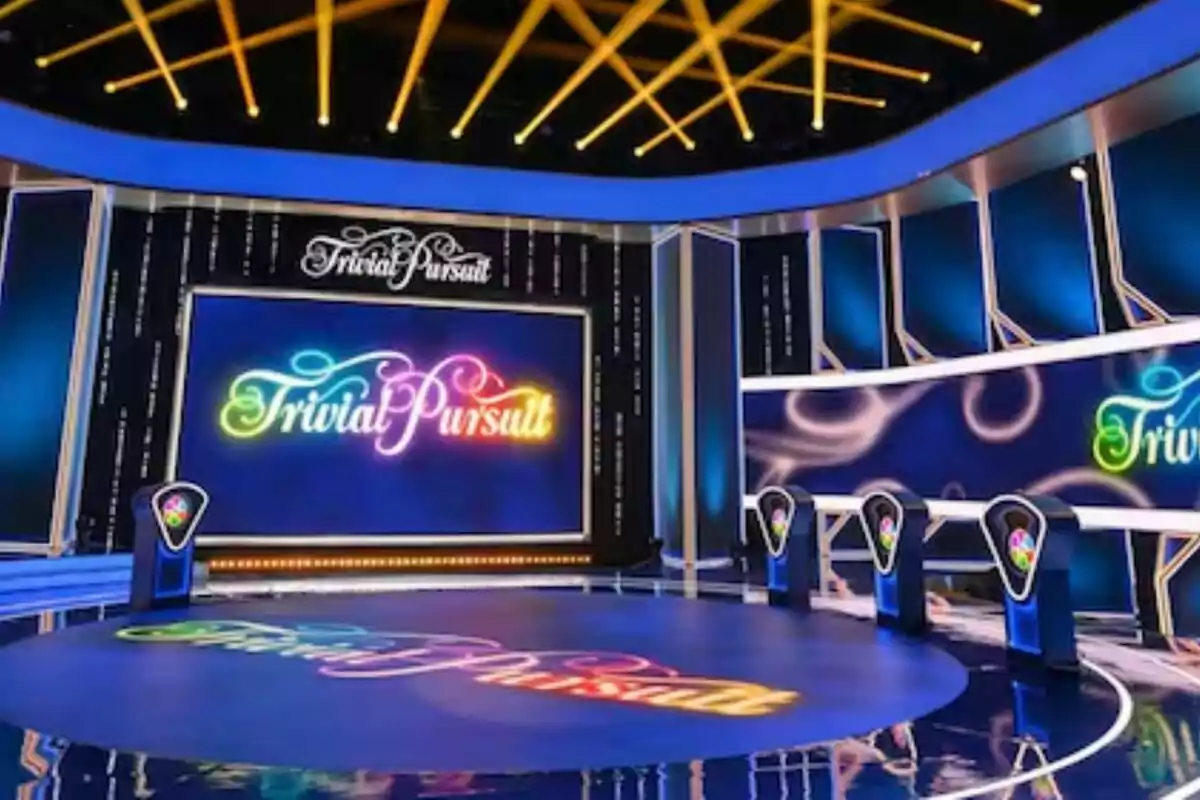 Escenario colorido de un programa de concursos con el logo de Trivial Pursuit en pantallas grandes y podios iluminados para los participantes
