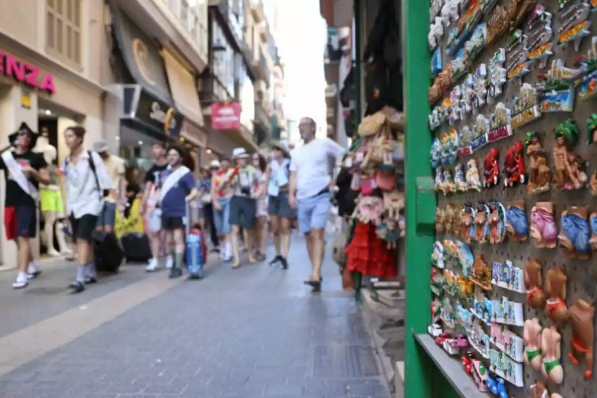 Un grupo de personas camina por una calle comercial llena de tiendas, mientras en primer plano se ven imanes de recuerdo en un puesto.