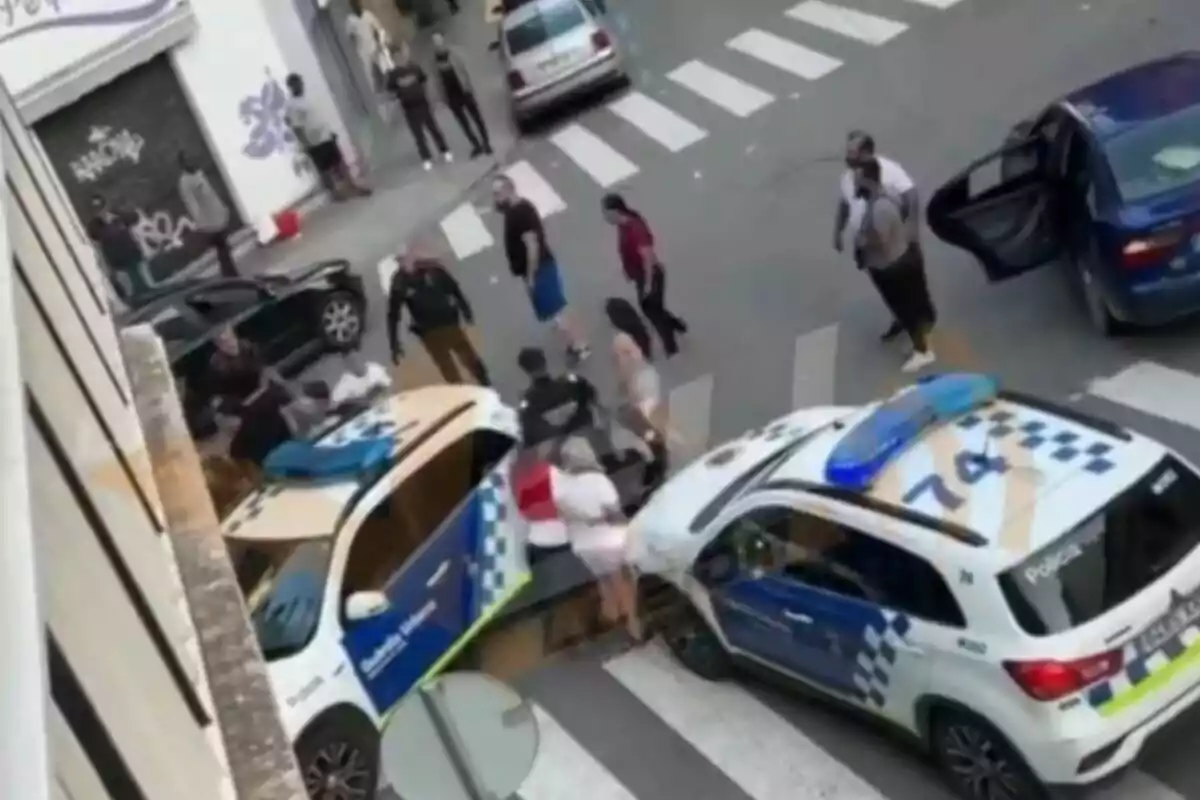 En la imagen se observa una escena urbana con dos vehículos policiales y varias personas alrededor, algunos de ellos parecen estar interactuando con los agentes de policía.