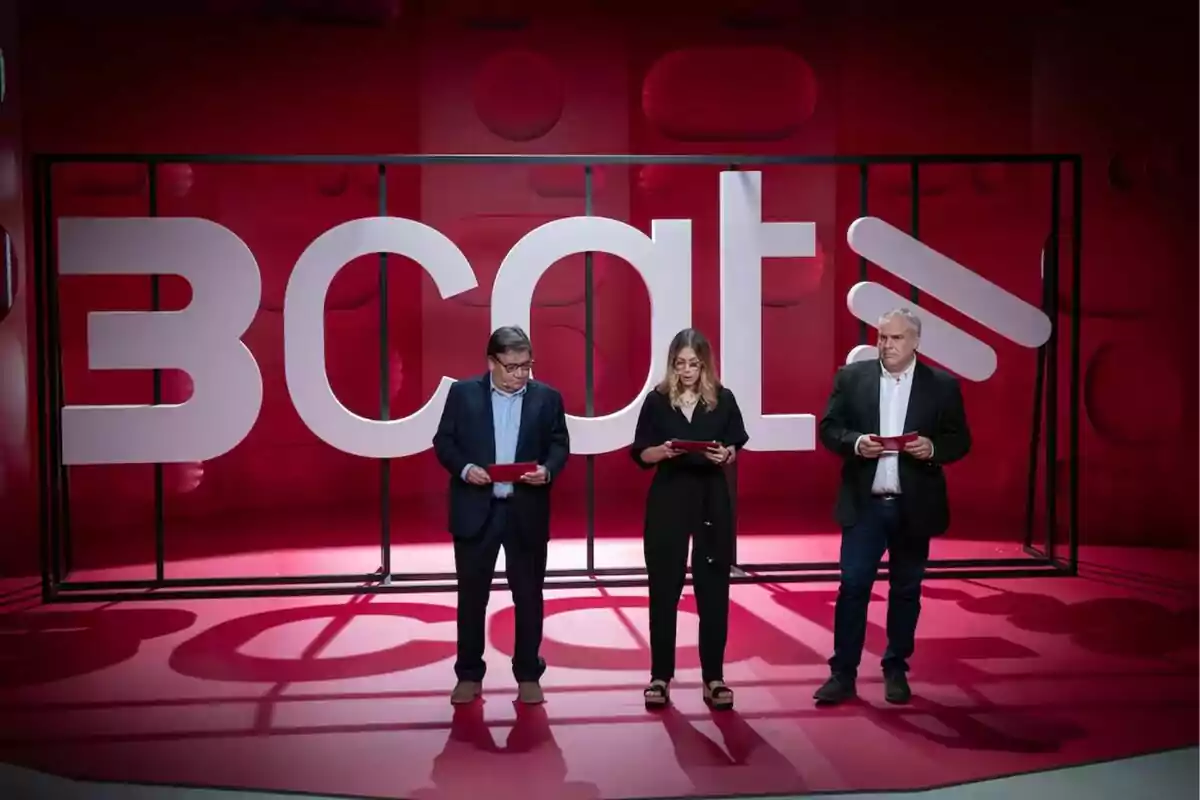 Tres personas de pie en un set de televisión con fondo rojo y las letras BCAT en grande detrás de ellos