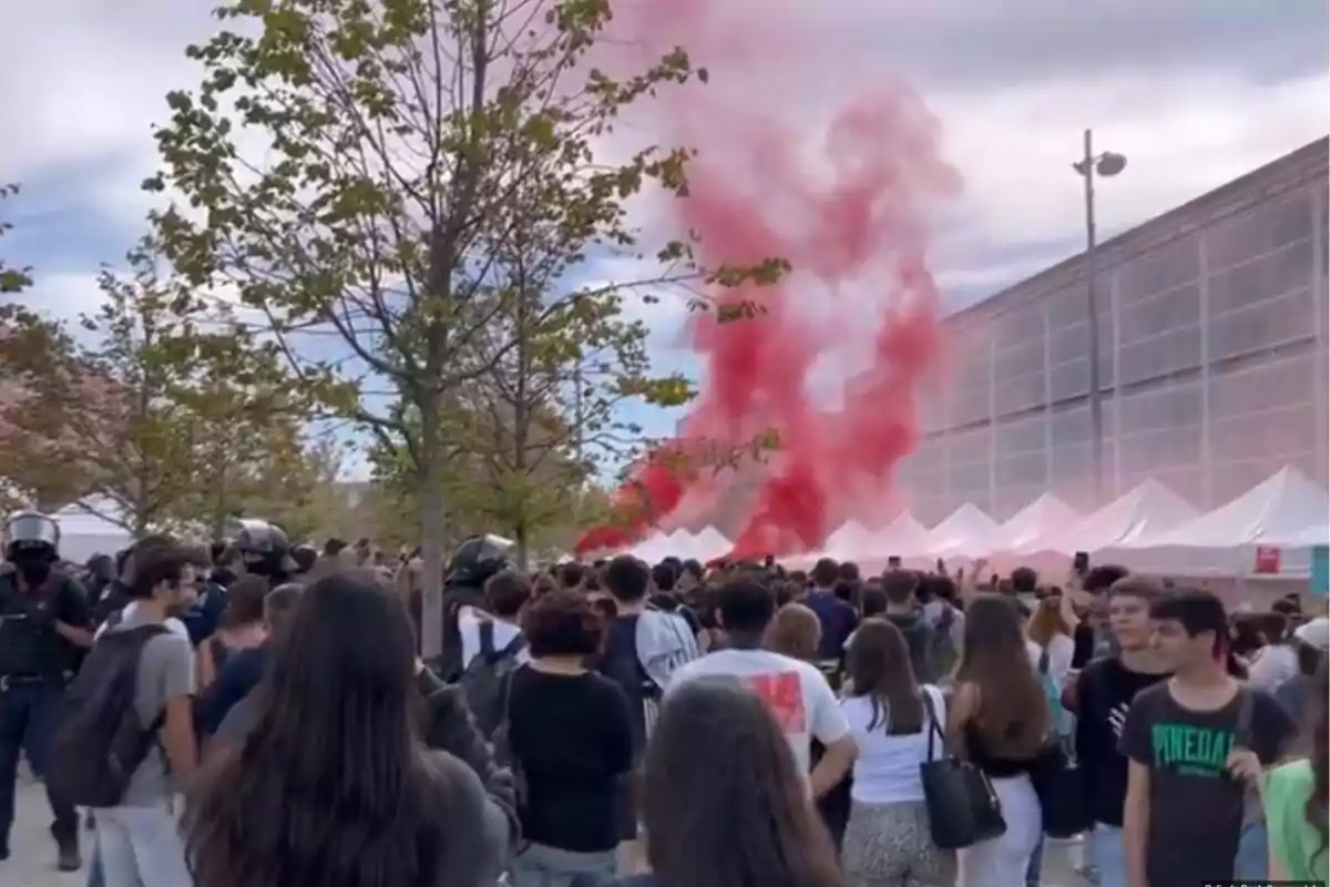 Una multitud de personas se reúne al aire libre, con humo rojo elevándose en el fondo y varias carpas blancas alineadas a lo largo de un edificio.