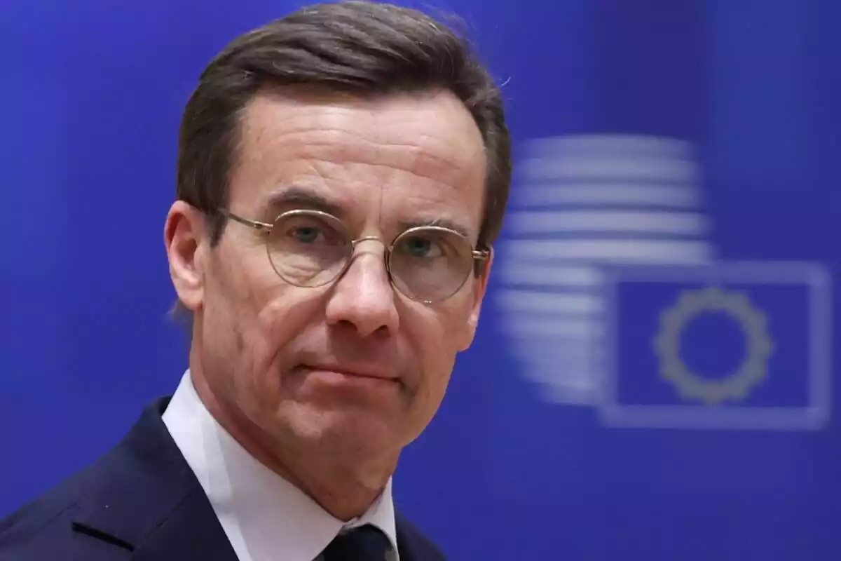 Primer plano de Ulf Kristersson, con gafas, mirando a cámara y con un fondo azul con una bandera europea difuminada