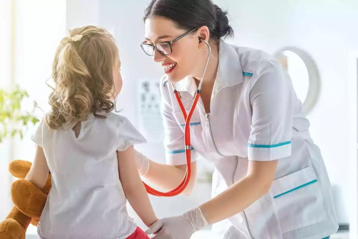 Una médica auscultando a una niña en un hospital, que sostiene un peluche en su mano Una médica auscultando a una niña en un hospital, que sostiene un peluche en su mano