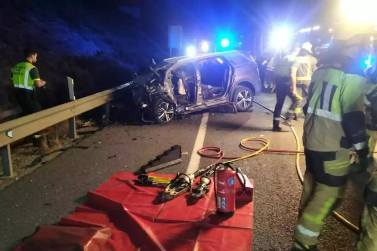 Accidente de tráfico nocturno con un coche gravemente dañado junto a la valla de seguridad, bomberos y equipo de rescate trabajando en la escena con herramientas y extintor en primer plano