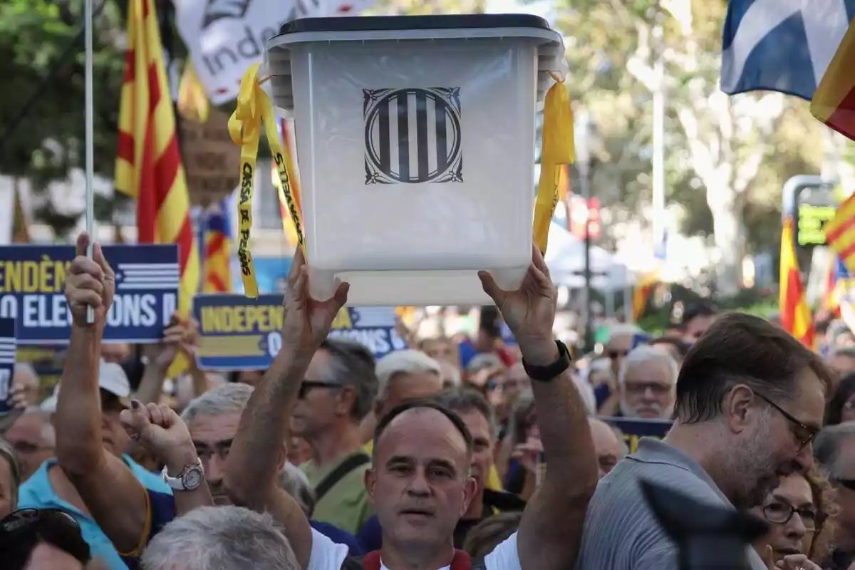 Un hombre calvo sujeta y levanta con las dos manos una urna de las que se usaron para el referéndum del 1 de octubre en Cataluña. Lo hace en medio de una manifestación, rodeado de decenas de personas