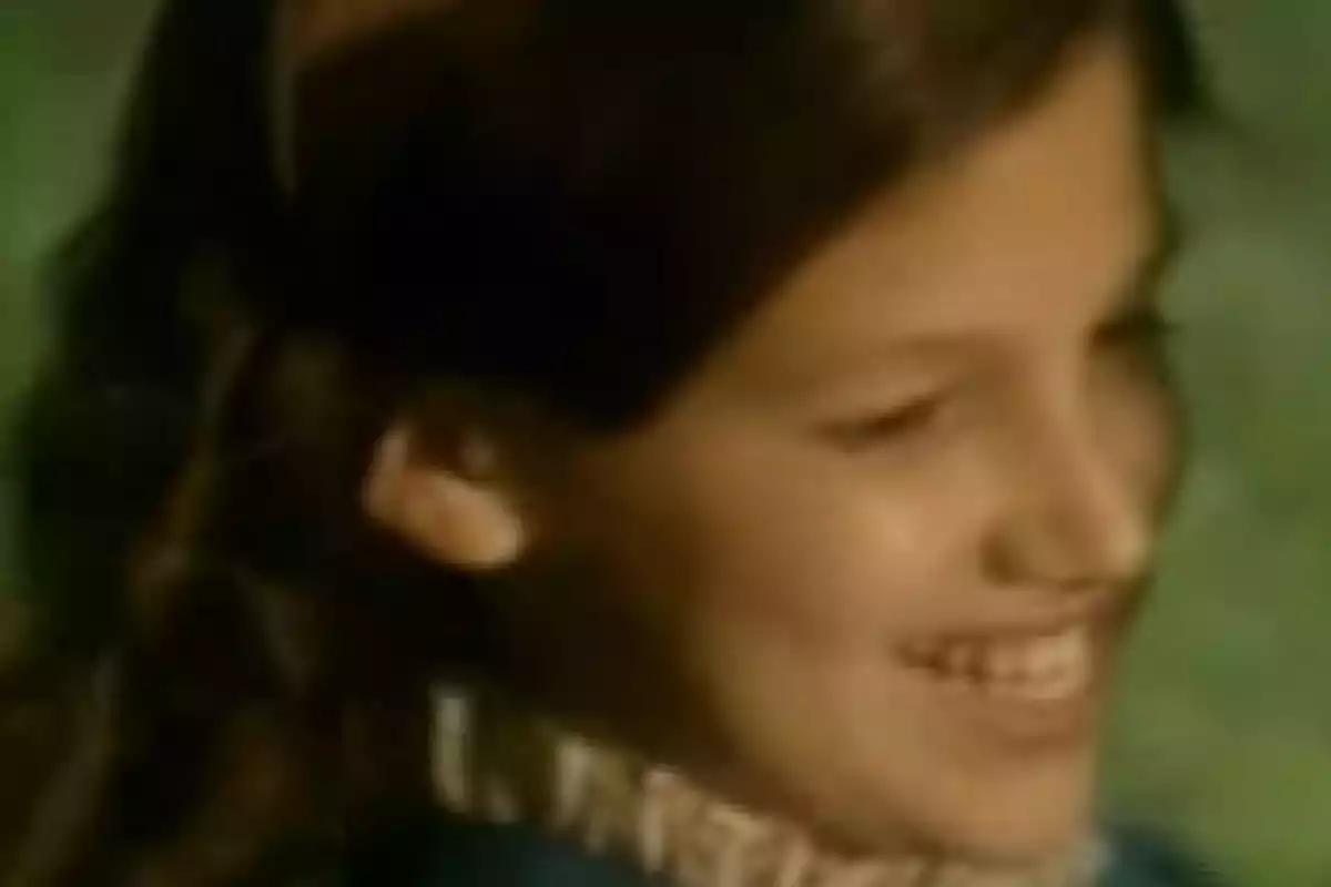 Una niña sonriente con cabello largo y oscuro, vestida con un suéter azul y una bufanda blanca, en un entorno verde. Una niña sonriente con cabello largo y oscuro, vestida con un suéter azul y una bufanda blanca, en un entorno verde.