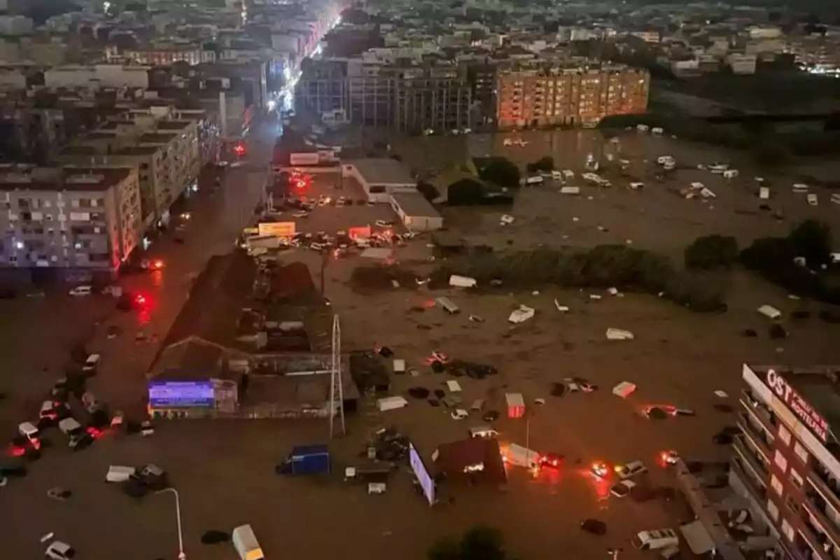 Vista aérea de una ciudad inundada durante la noche con calles cubiertas de agua y vehículos sumergidos, luces de emergencia reflejadas en el agua.