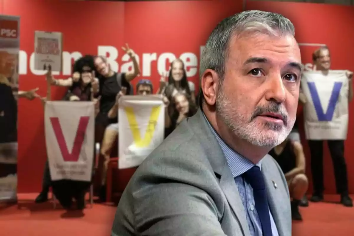 Hombre de cabello canoso y barba con traje y corbata en primer plano, al fondo personas sostienen pancartas con letras grandes y coloridas sobre un fondo rojo