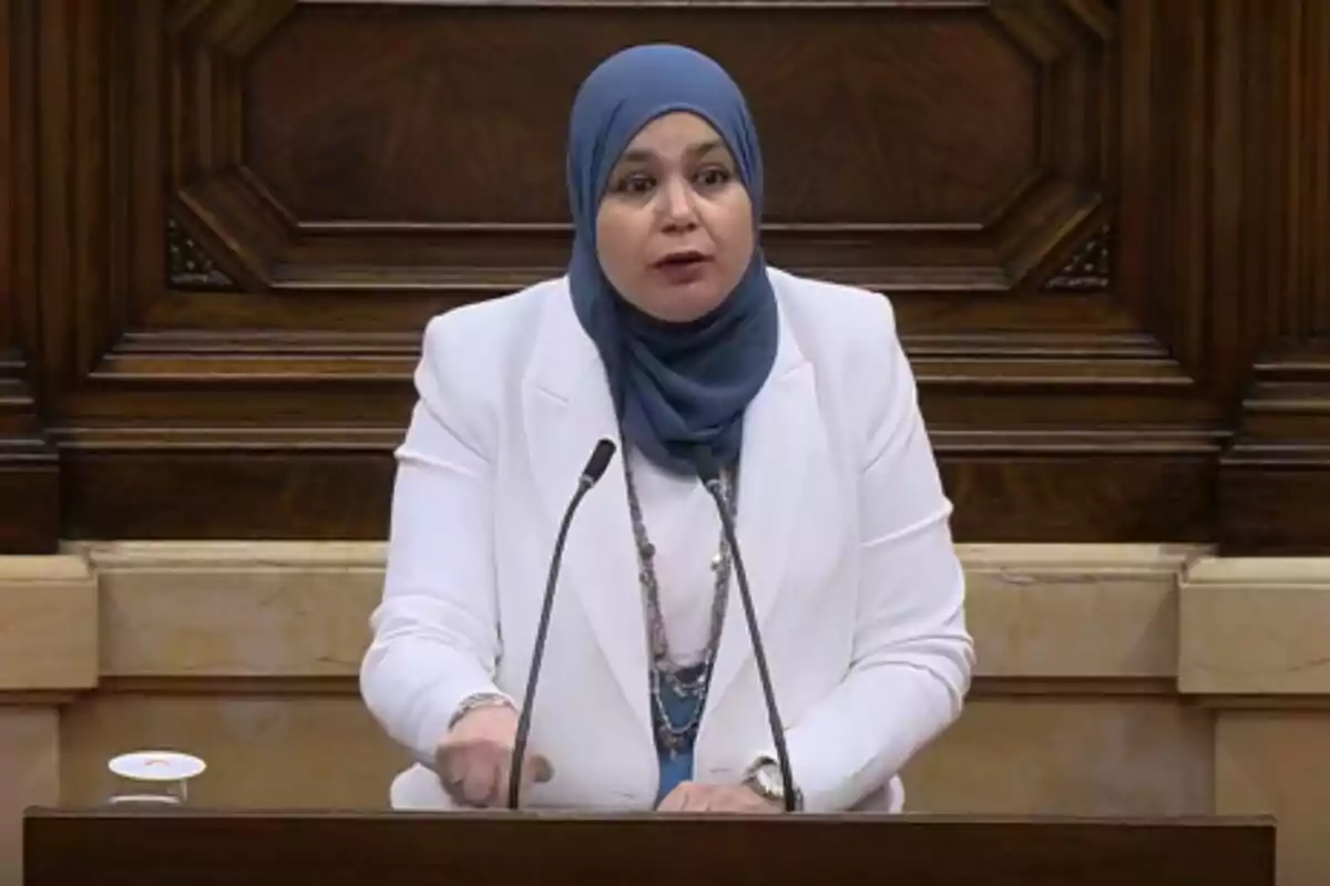 Una mujer con hiyab azul y traje blanco habla en un podio con micrófonos frente a un fondo de madera.