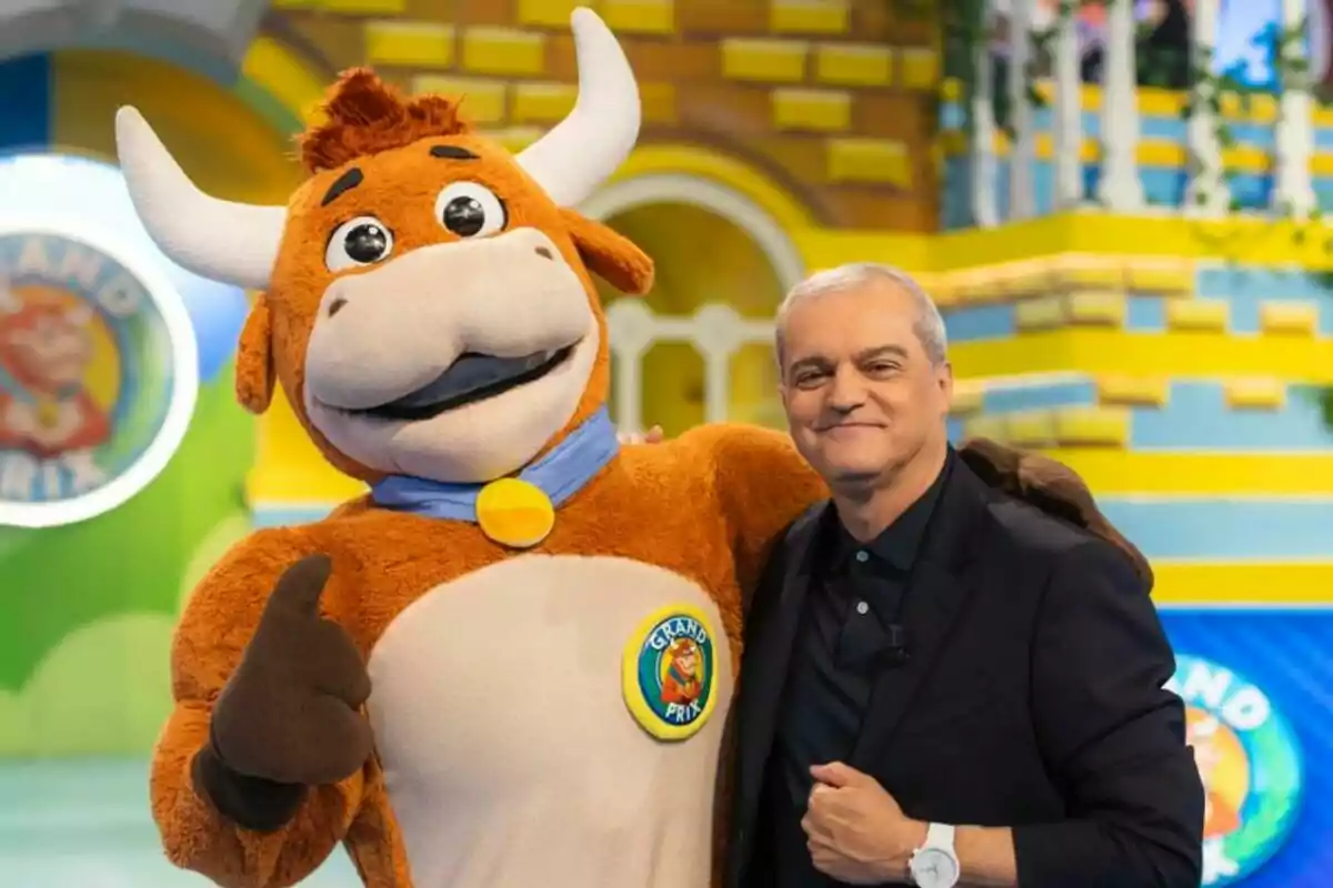 Ramón García sonriente posa junto a una persona disfrazada de vaquilla en un colorido set de El Grand Prix