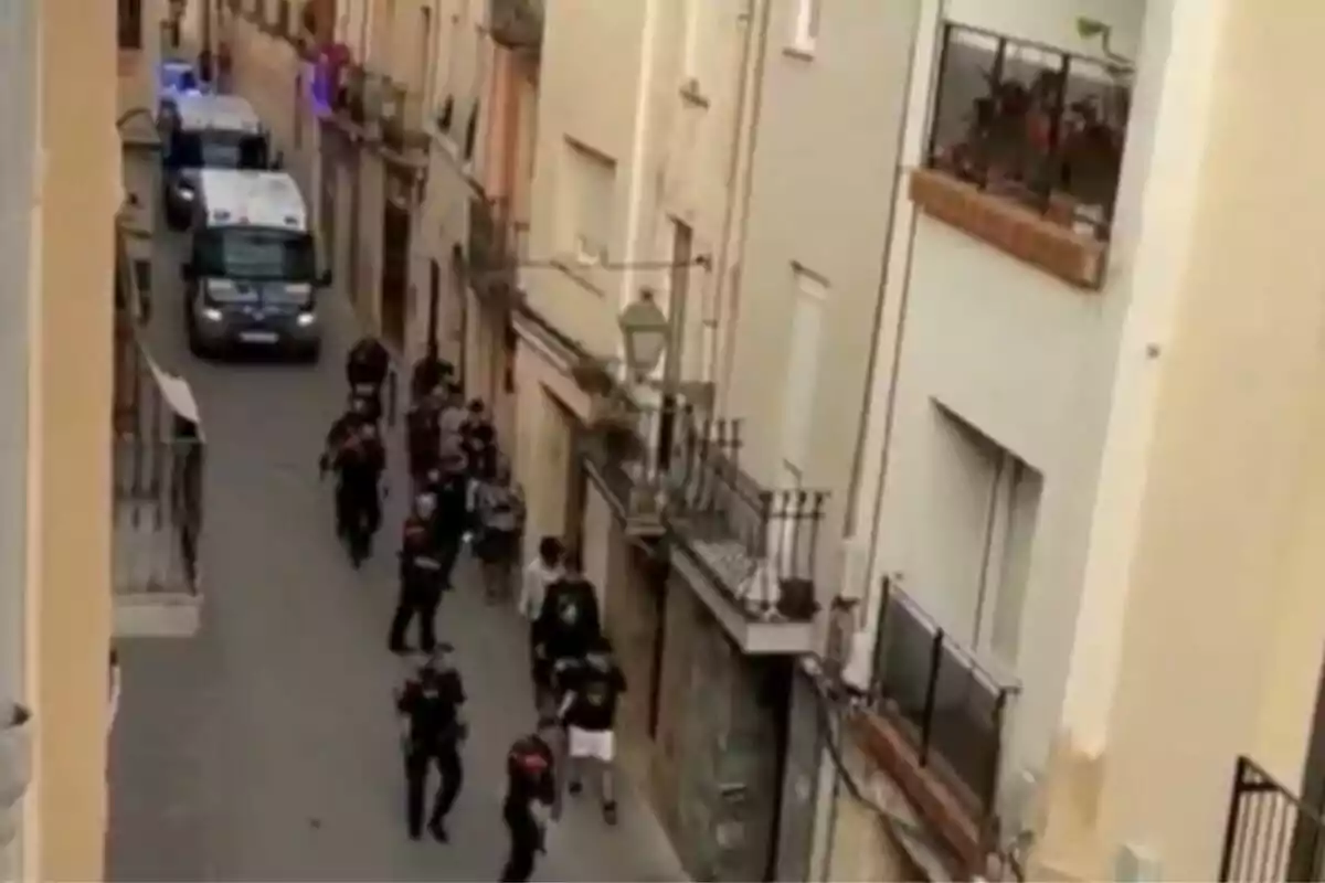 Un grupo de personas camina por una calle estrecha flanqueada por edificios altos, mientras varios vehículos policiales están estacionados al fondo.