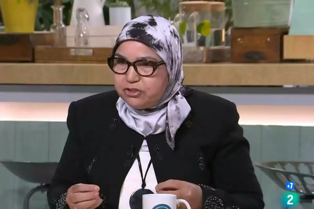 Una persona con gafas y un pañuelo en la cabeza está hablando en un entorno que parece ser un estudio de televisión o una cocina, con varios objetos y plantas en el fondo.