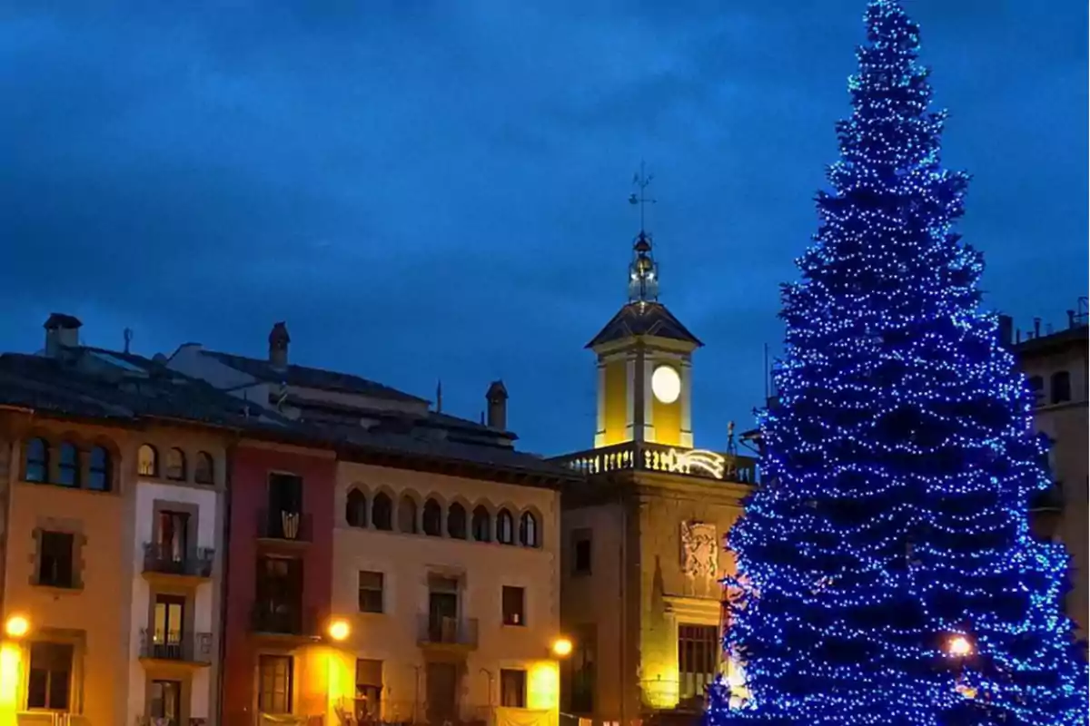 Una plaza iluminada con un gran árbol de Navidad decorado con luces azules y un edificio con un reloj en la torre al fondo.