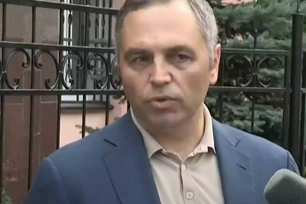 Un hombre con chaqueta azul y camisa beige habla frente a una reja metálica mientras sostiene un micrófono. Un hombre con chaqueta azul y camisa beige habla frente a una reja metálica mientras sostiene un micrófono.