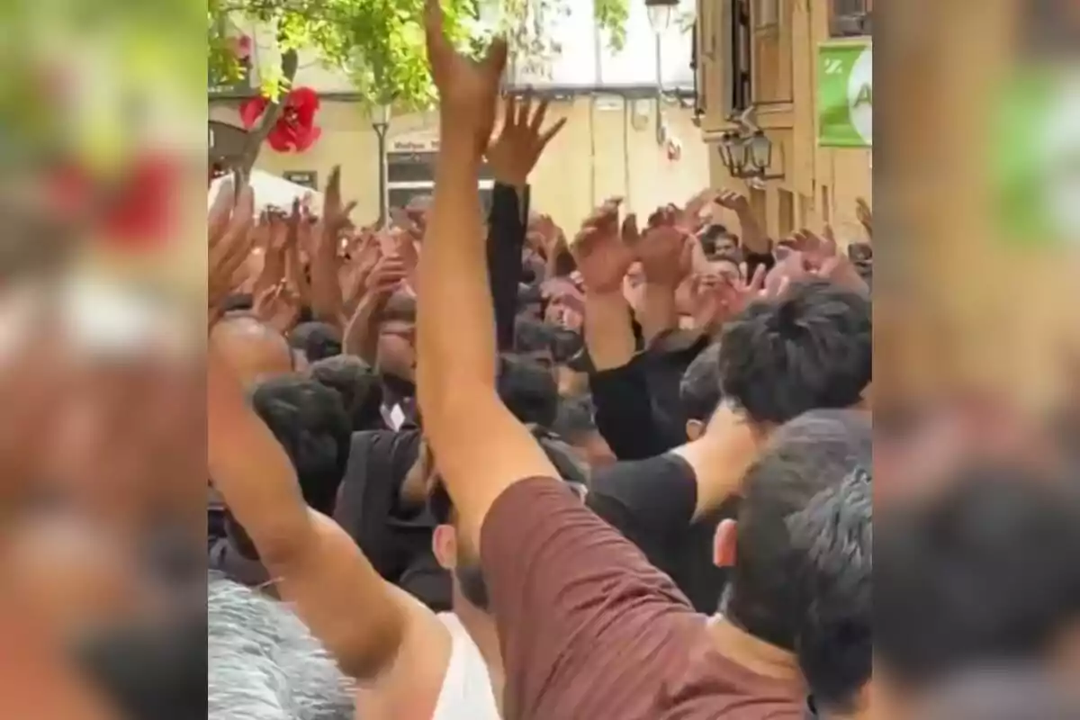 Una multitud de personas con los brazos levantados en una calle, rodeada de edificios y árboles.