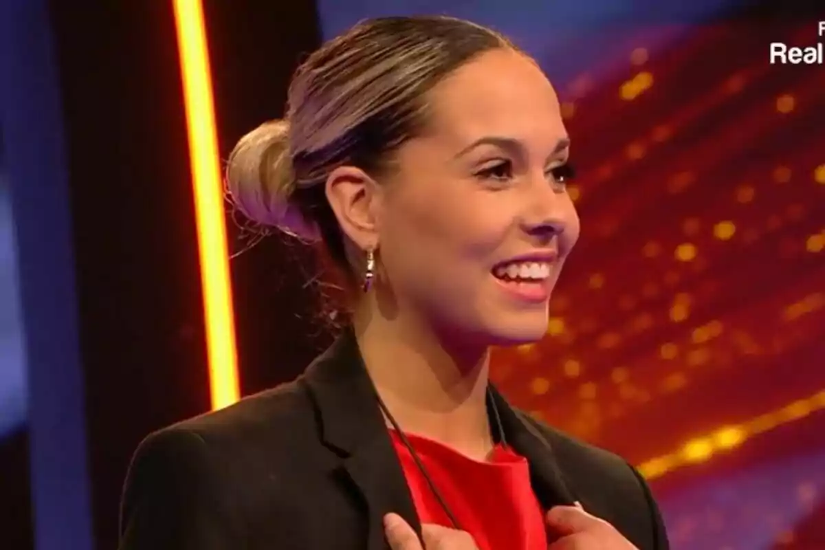 Violeta sonriente con el cabello recogido y vestida con un blazer negro y blusa roja en un set iluminado en Gran Hermano.