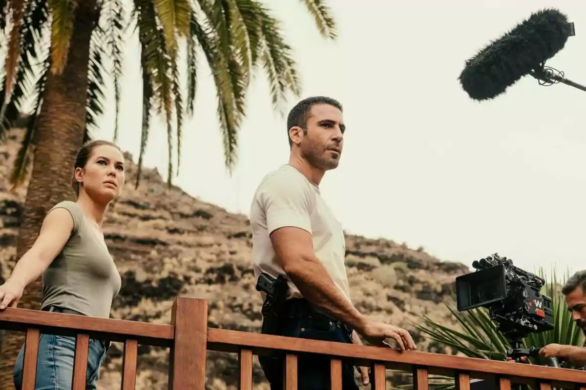 Fotografía de Miguel Ángel Silvestre y Katia Fellin de pie junto a una barandilla de madera, con una cámara de filmación y un micrófono de boom en el fondo, en un entorno al aire libre con palmeras y colinas en el rodaje de Weiss & Morales