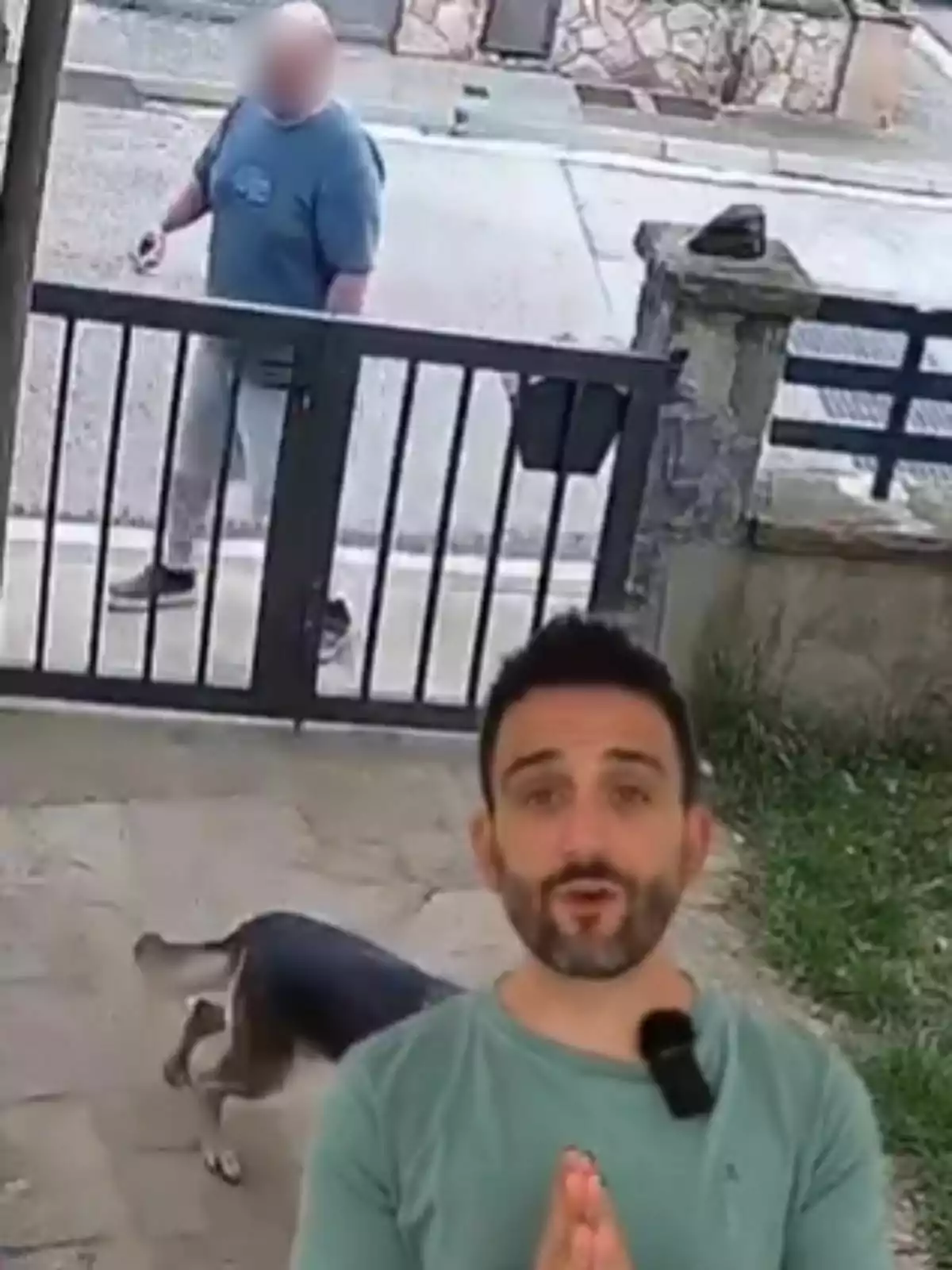 Un hombre con camiseta azul camina por la acera mientras un perro está en el patio detrás de una reja y un hombre con camiseta verde aparece en primer plano. Un hombre con camiseta azul camina por la acera mientras un perro está en el patio detrás de una reja y un hombre con camiseta verde aparece en primer plano.