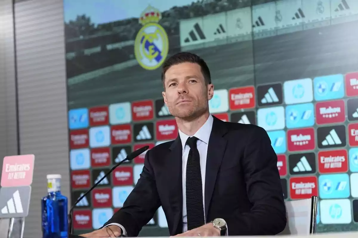 Hombre con traje oscuro sentado en una conferencia de prensa con el escudo del Real Madrid y un fondo de patrocinadores detrás Hombre con traje oscuro sentado en una conferencia de prensa con el escudo del Real Madrid y un fondo de patrocinadores detrás