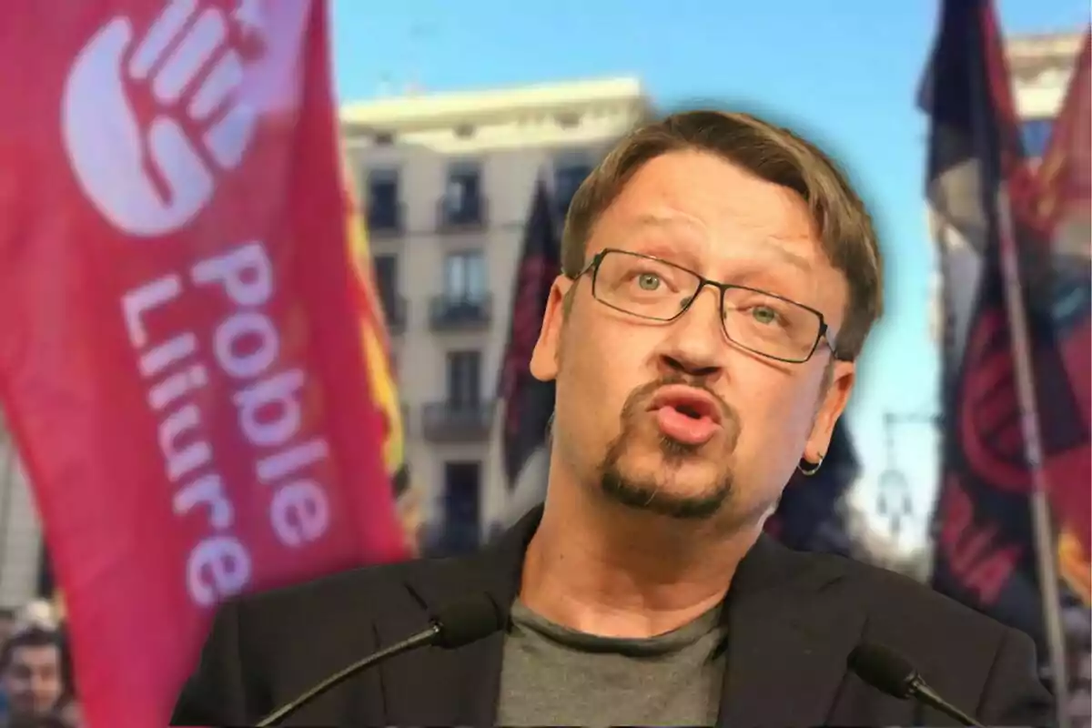 Un hombre con gafas y barba hablando frente a un micrófono con banderas de fondo.