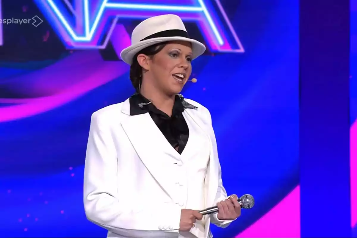 Yenesi vestida con traje blanco y sombrero sosteniendo un micrófono en un escenario con fondo azul y rosa de Tu Cara Me Suena Yenesi vestida con traje blanco y sombrero sosteniendo un micrófono en un escenario con fondo azul y rosa de Tu Cara Me Suena