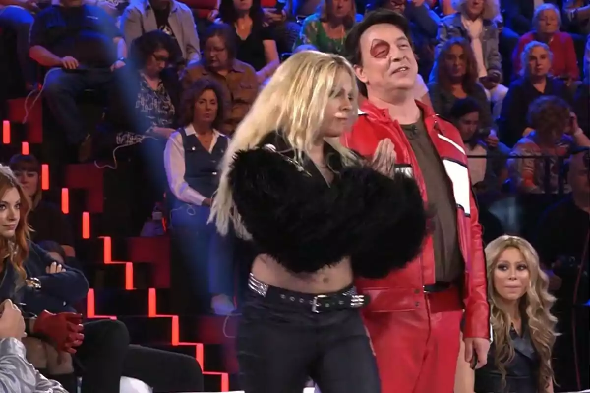 Un grupo de personas está sentado en un estudio de televisión, mientras dos individuos en primer plano, Goyo Jiménez con chaqueta roja y Yenesi con chaqueta negra de peluche, parecen estar participando en Tu Cara Me Suena. Un grupo de personas está sentado en un estudio de televisión, mientras dos individuos en primer plano, Goyo Jiménez con chaqueta roja y Yenesi con chaqueta negra de peluche, parecen estar participando en Tu Cara Me Suena.